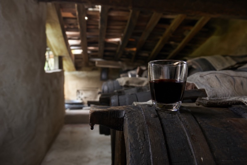 A small glass of dark syrupy balsamic on a barrel stave with a drip at the spout in a Modena attic