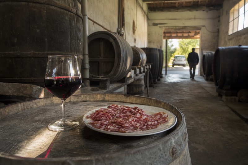 A glass of deep ruby Lambrusco and sliced salumi on a weathered barrel top in a Nonantola cellar