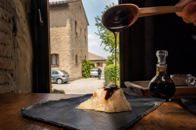 A wooden spoon drizzling thick balsamic over a Parmigiano shard on a slate board in a Modena acetaia