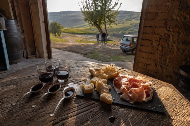 Three glasses of Lambrusco and tiny spoons of dark balsamic on a wooden table in a Castelvetro di Modena cellar