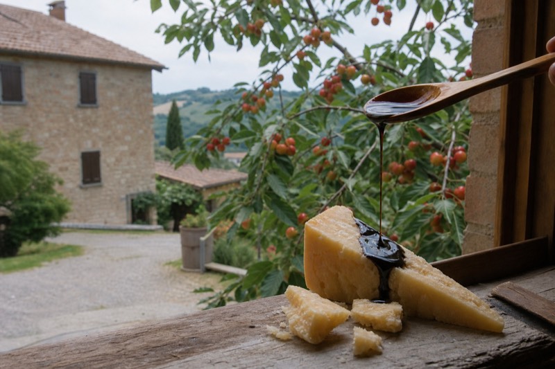 A wooden spoon drizzles balsamic onto Parmigiano while cherry branches press against a stone window in Vignola