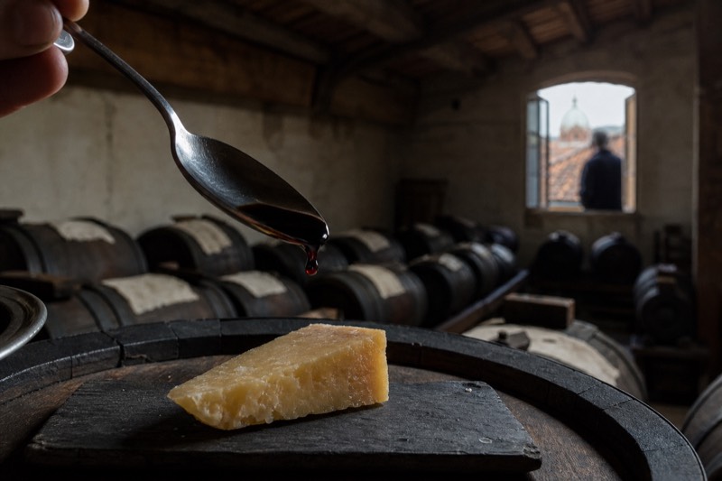 A silver spoon holding a syrupy drop of aged balsamic beside Parmigiano on a slate board in a dim Modena attic