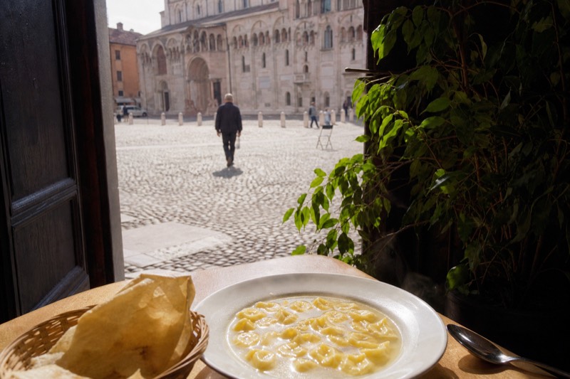 A steaming bowl of tortellini in brodo on a table in Piazza Grande with Modena's cathedral blurred beyond a doorway