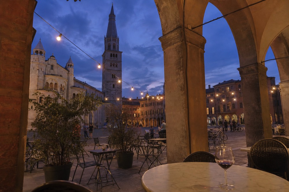 An evening piazza with stone buildings, arched walkways and cafe tables under lights