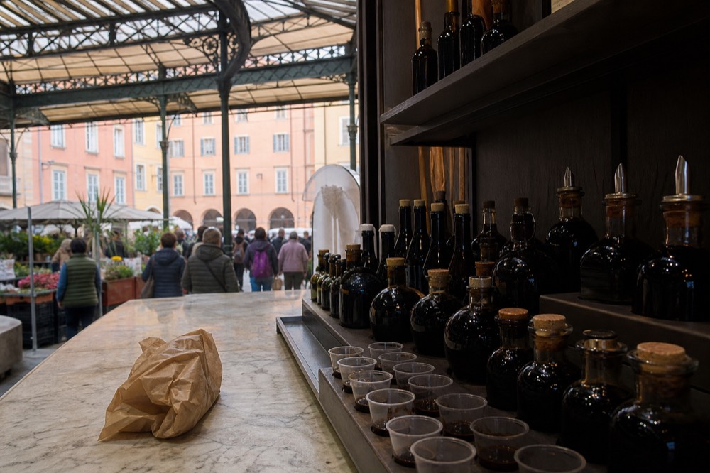 A market stall counter with shelves of balsamic bottles and tasting cups