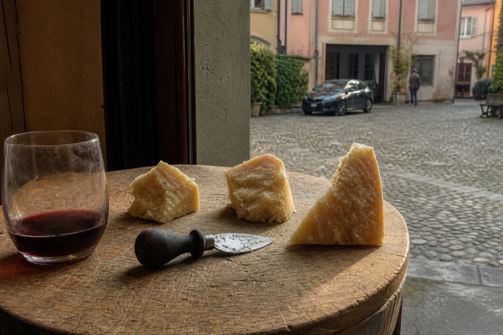 A wooden board with wedges of Parmigiano Reggiano at different ages beside a knife and a glass of red wine