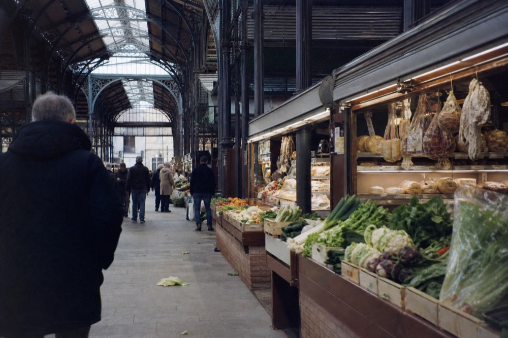 A covered market hall with stalls of vegetables, cheese and cured meats under a high glass roof