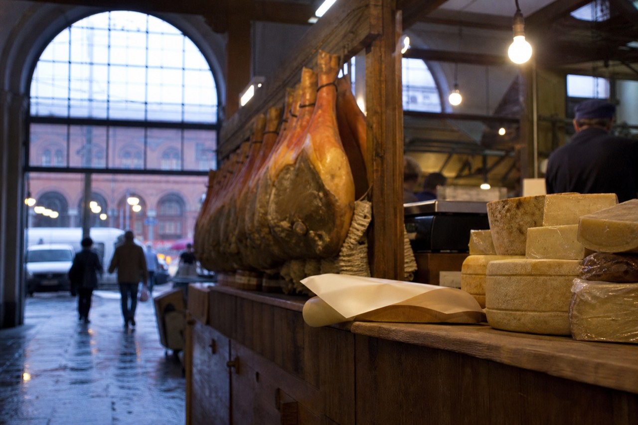 A wooden market counter stacked with whole legs of cured pork and wheels of aged cheese under warm hanging lights