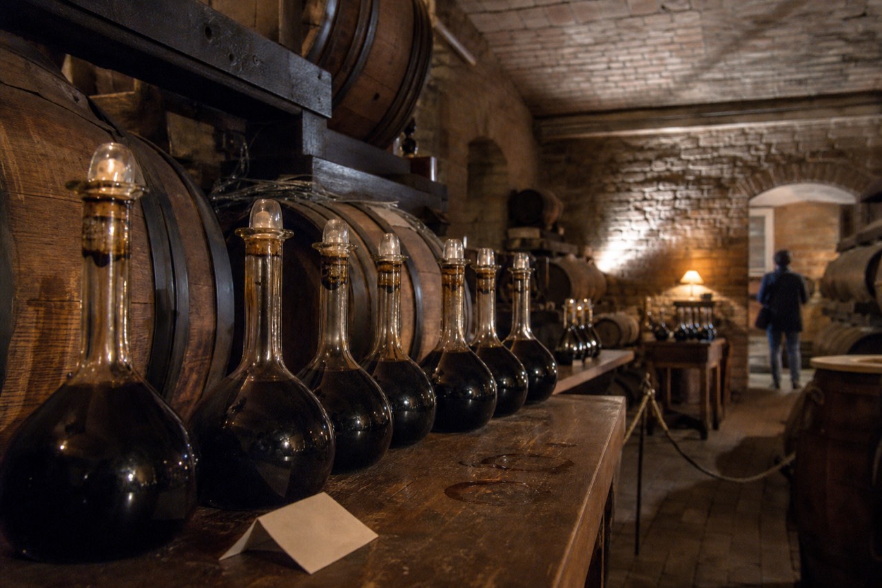 A row of small balsamic vinegar bottles and wooden barrels in a dim Modena cellar