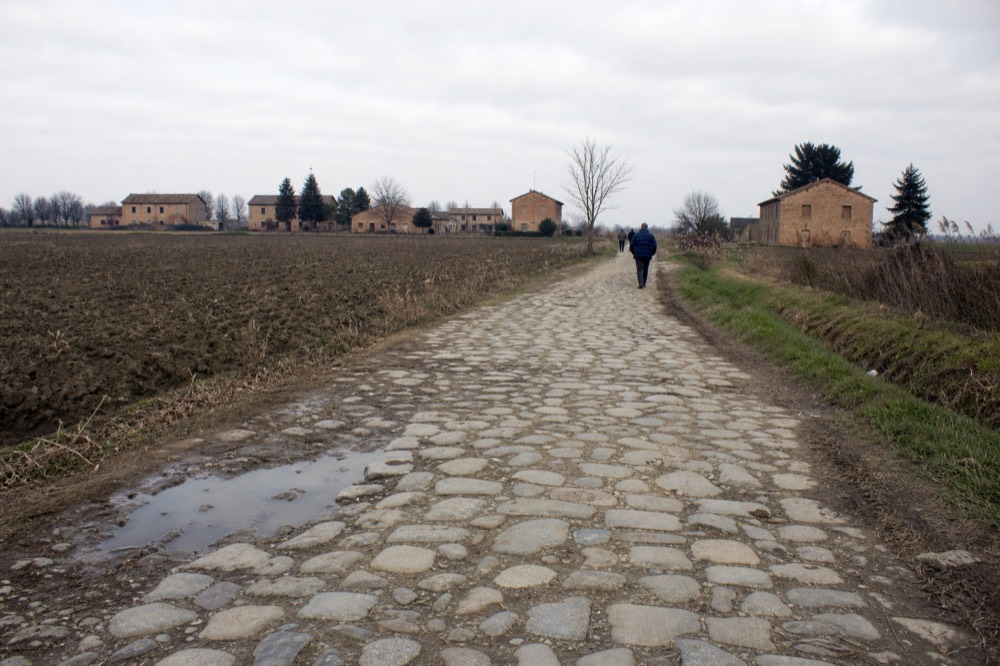 An old stone-paved road stretching straight through flat countryside with farm buildings on either side