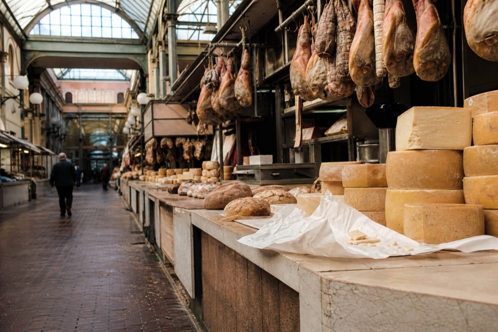 A market stall with stacked cheeses, cured hams and bread under a glass roof