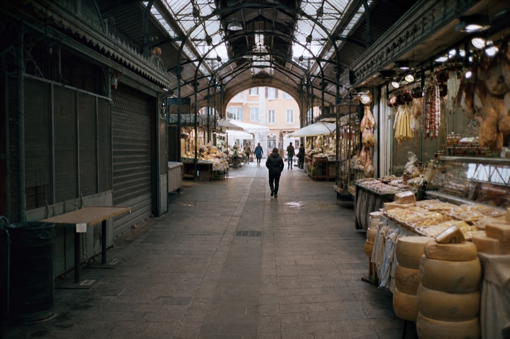 A covered market hall with stalls of cured meats, cheese wheels and fresh pasta under iron and glass