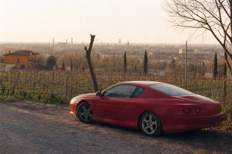 Ferrari and food category card showing a red sports car beside a vineyard, for full-day Modena tours pairing car museums with tastings
