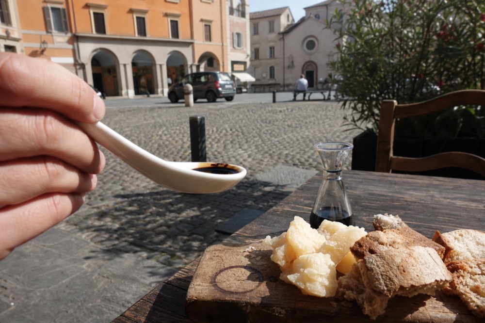 A ceramic spoon holding dark glossy balsamic vinegar above a tasting board