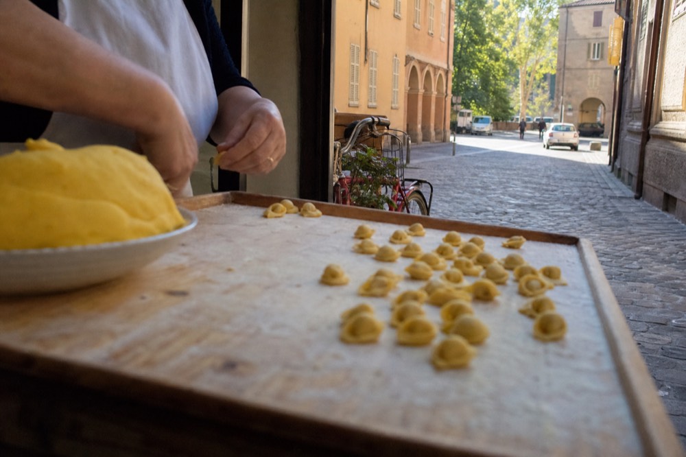 Hands folding small tortellini on a floured wooden table beside a bowl of egg dough