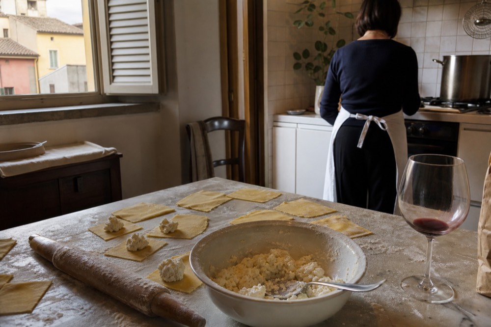 A flour-dusted kitchen counter with a rolling pin, cut pasta squares and a bowl of ricotta filling