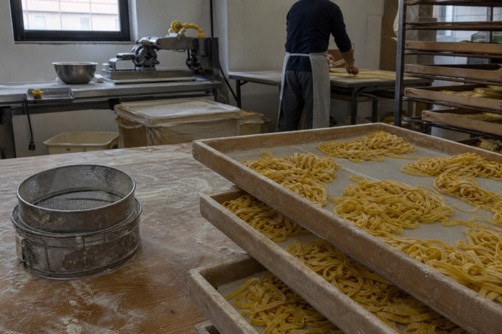 A pasta workshop bench with stacked trays of freshly cut tagliatelle and a flour sieve