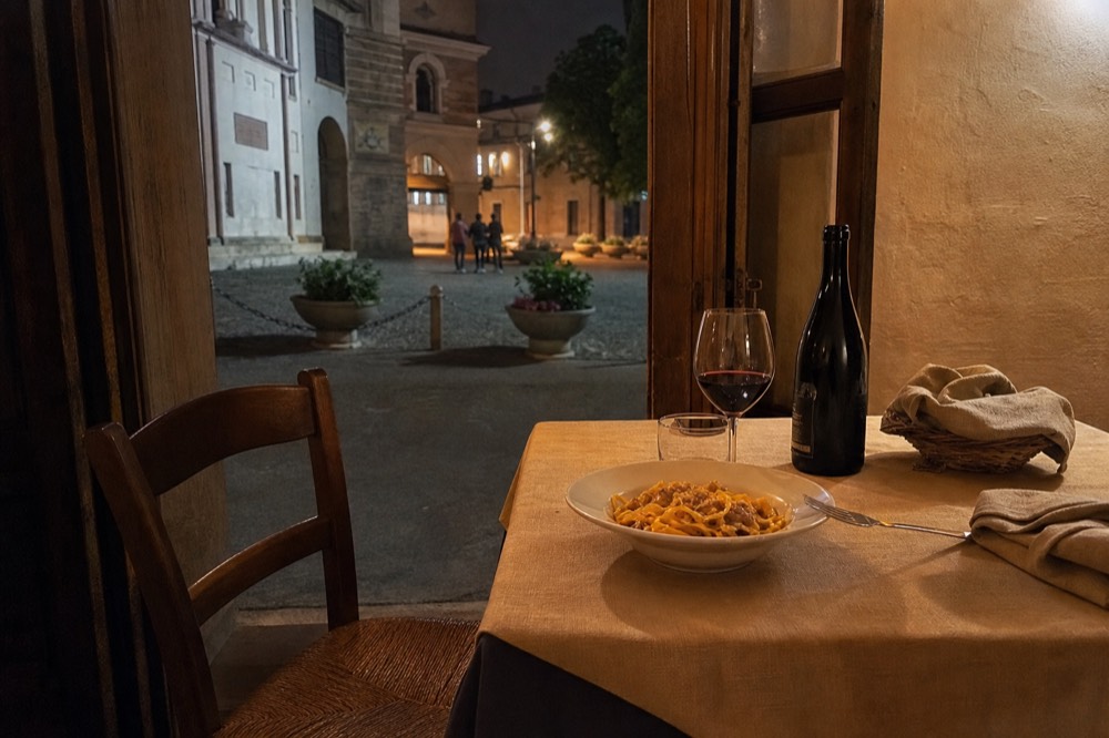 A laid table with a bowl of tagliatelle, a bottle of Lambrusco and folded napkins in a warm dining room