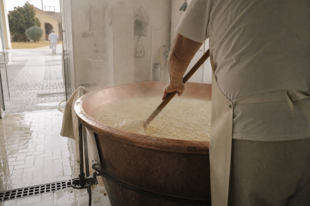 Copper cheese vat with steam rising as a worker stirs curd with a large paddle