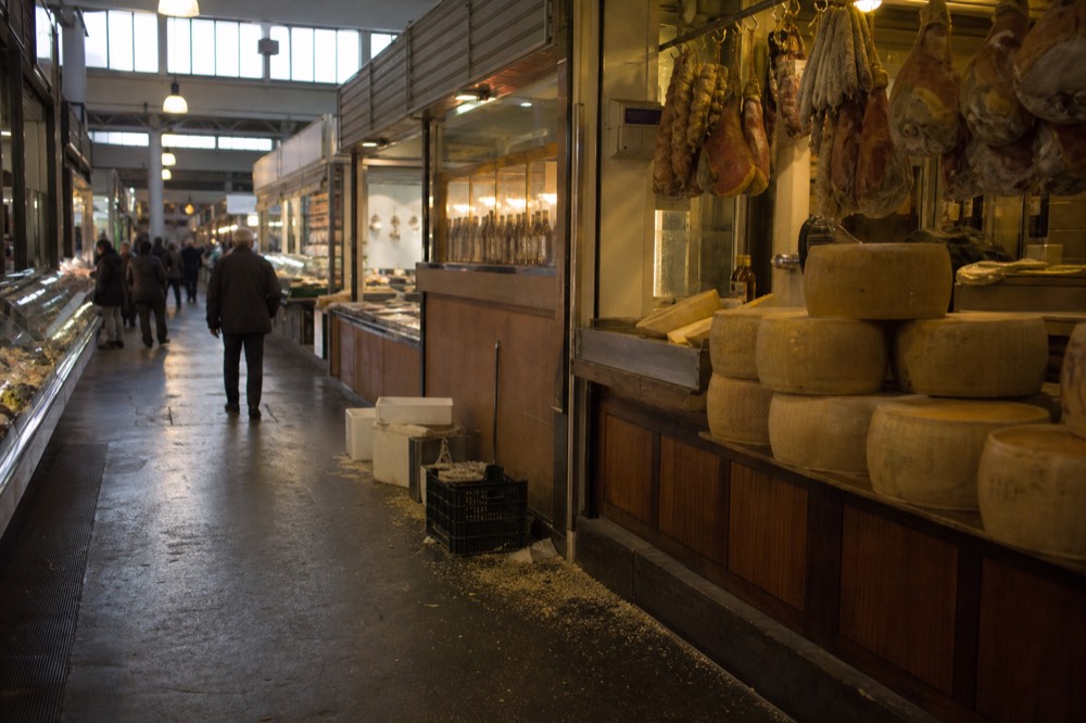 Market stalls displaying hanging cured meats and cheese wheels under warm lights