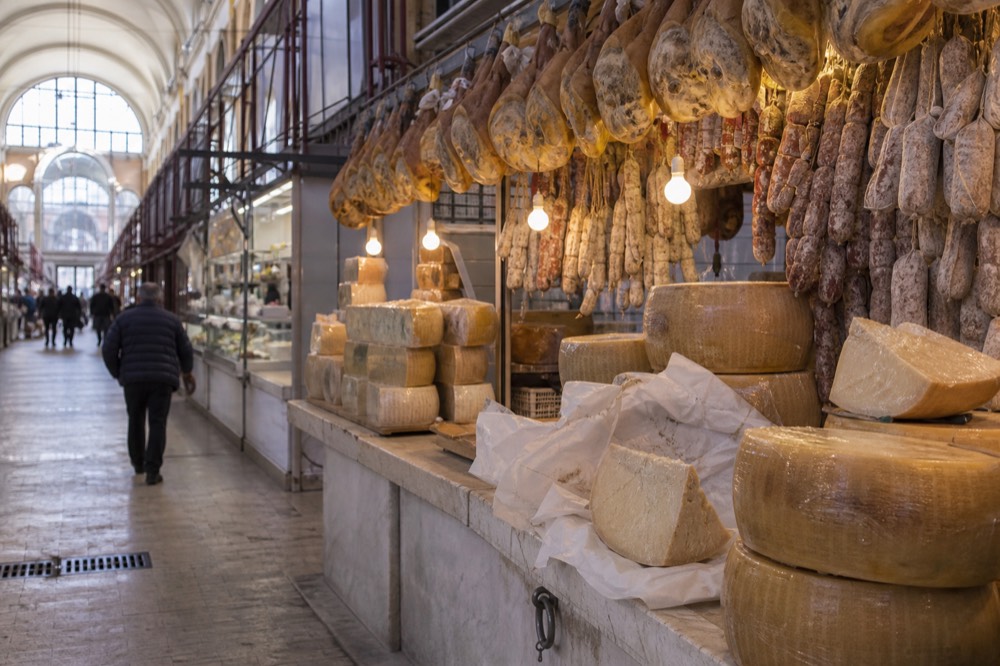 Market stalls with hanging cured meats and wheels of cheese under warm lights