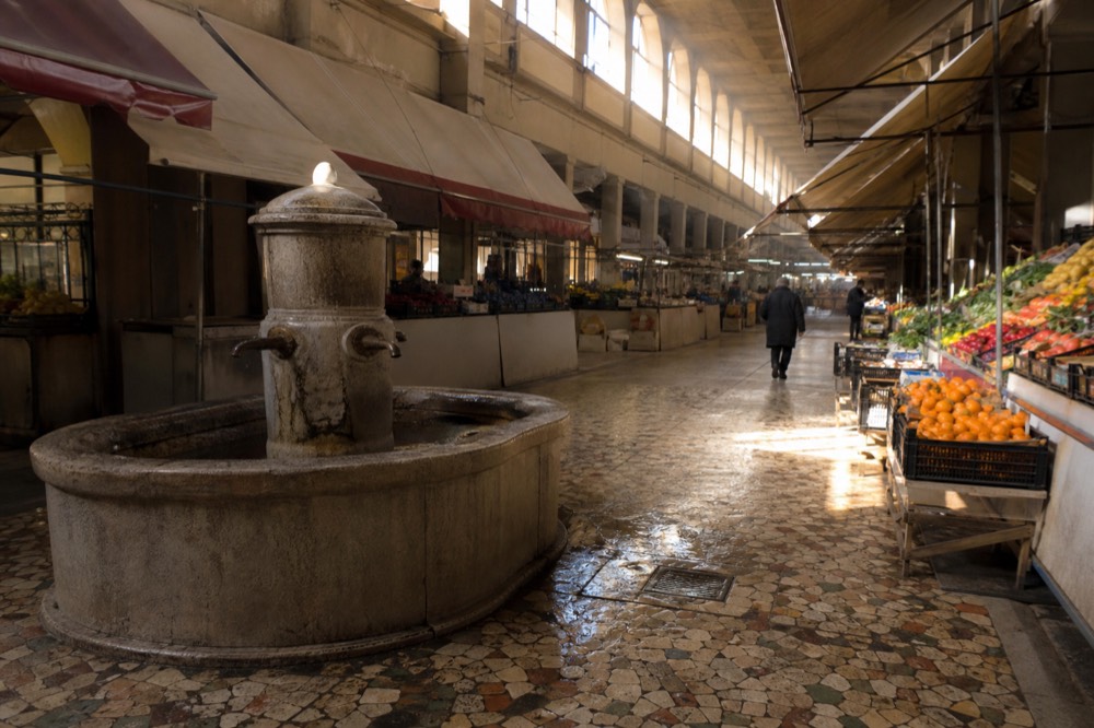 A covered market aisle with a stone fountain and stall awnings in morning light