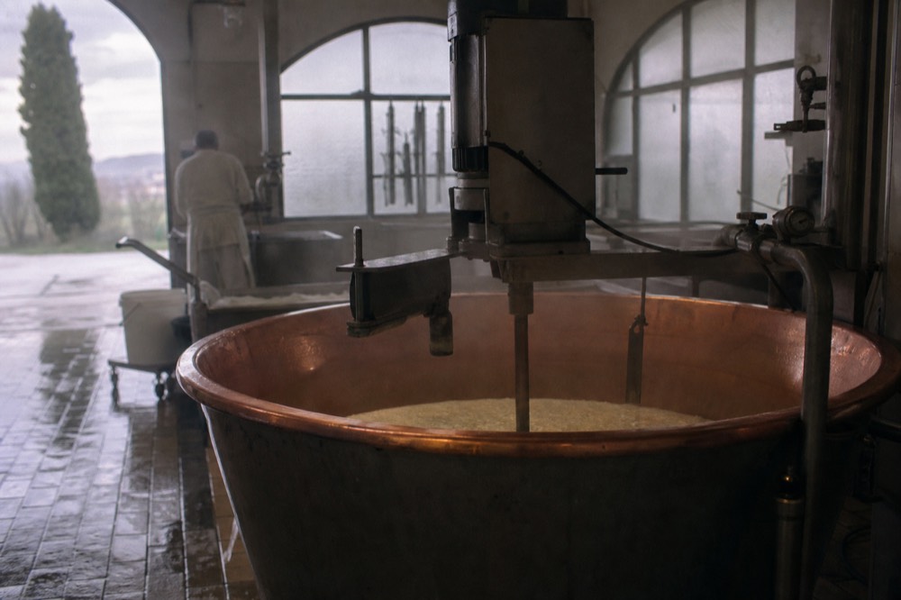 A copper cheesemaking vat and steaming whey inside a working dairy room