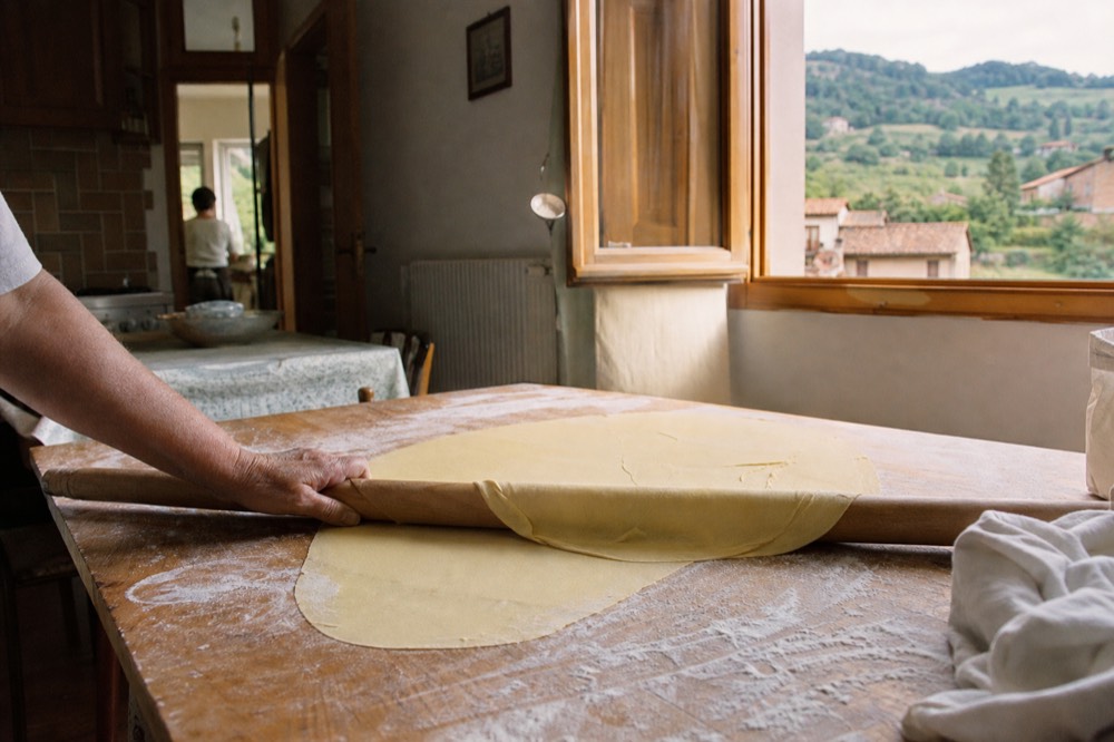 Fresh egg pasta dough being rolled thin with a wooden rolling pin on a floured board