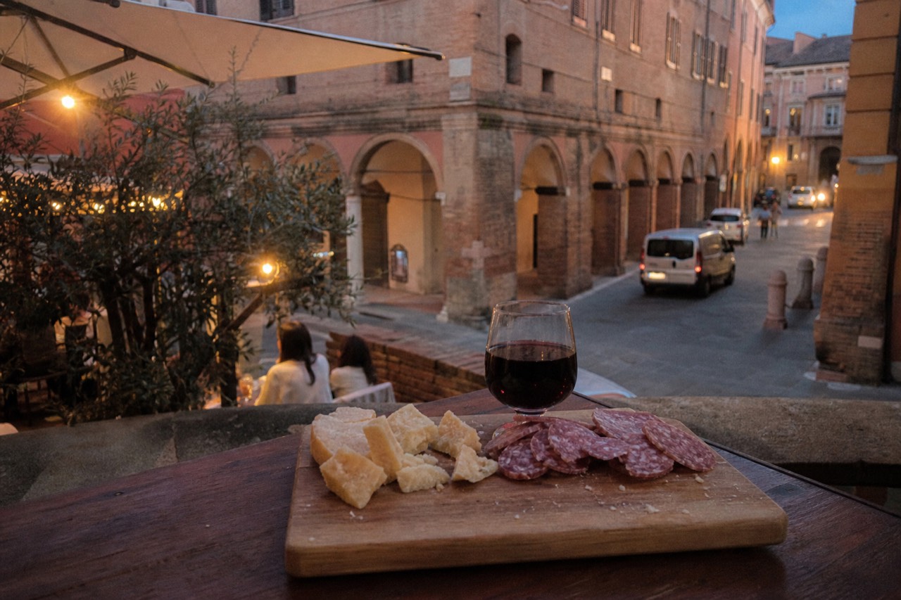 A wooden tasting board with Parmigiano Reggiano chunks, sliced salami and a small glass of red wine on a rustic table