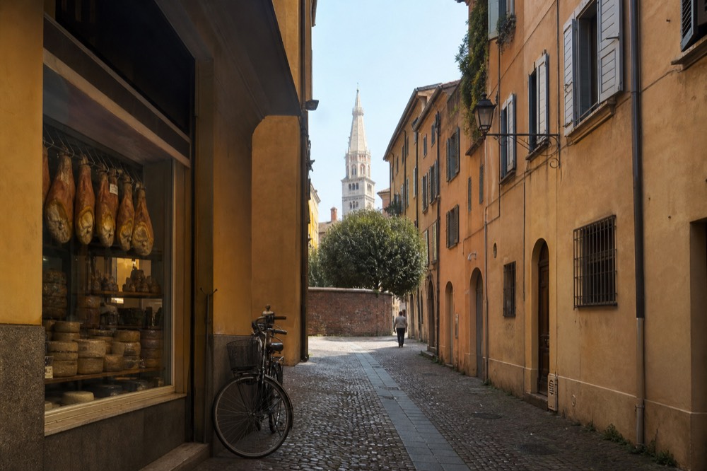 A narrow Modena street with a traditional salumeria window filled with hanging prosciutto and cheese wheels