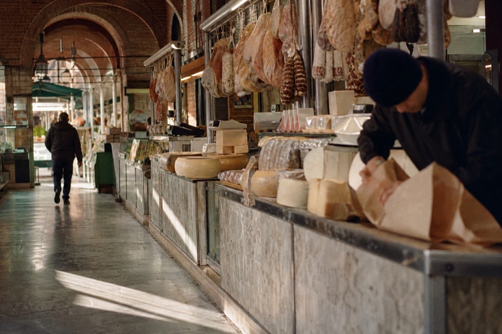 Market stalls with hanging cheeses and cured meats under the arches of a covered hall