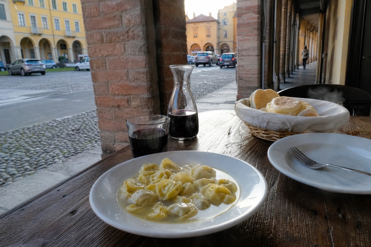 A rustic wooden table set with a bowl of tortellini in broth, a basket of tigelle and a bottle of Lambrusco