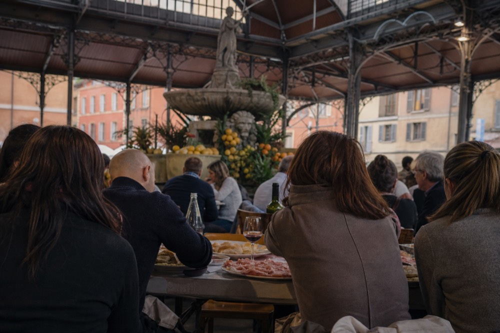 Diners sitting at a communal table inside a covered market, plates of cured meats and bread in front of them