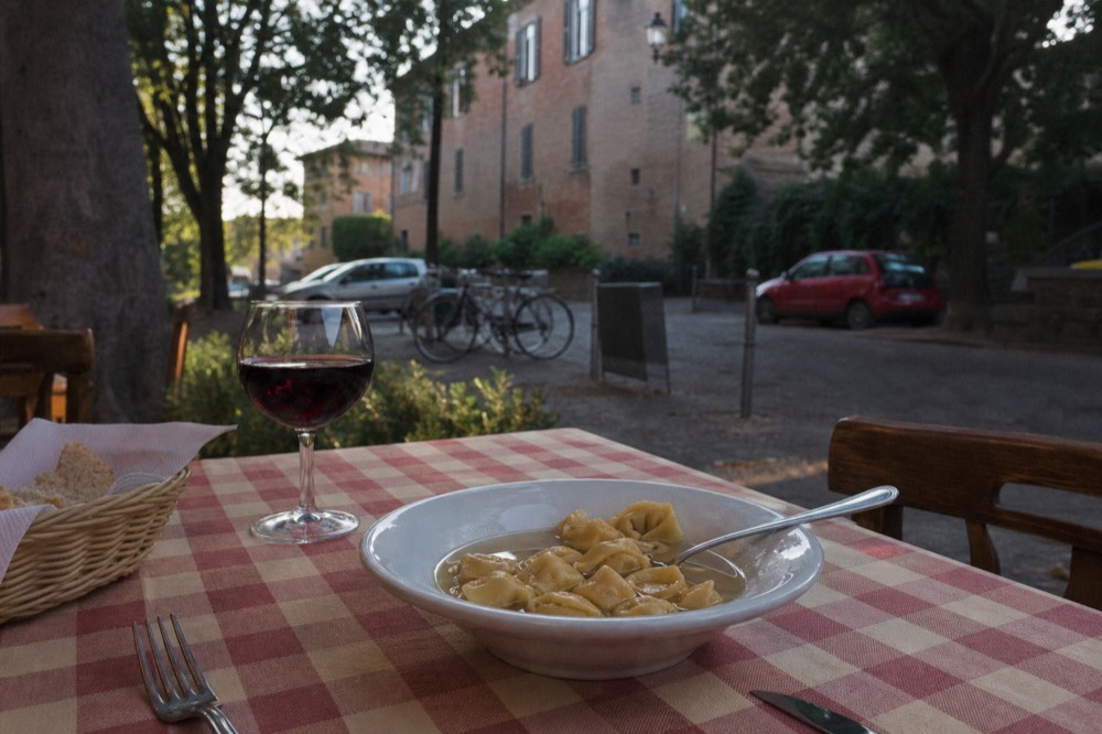 A bowl of tortellini in brodo with a spoon and a glass of red wine beside it