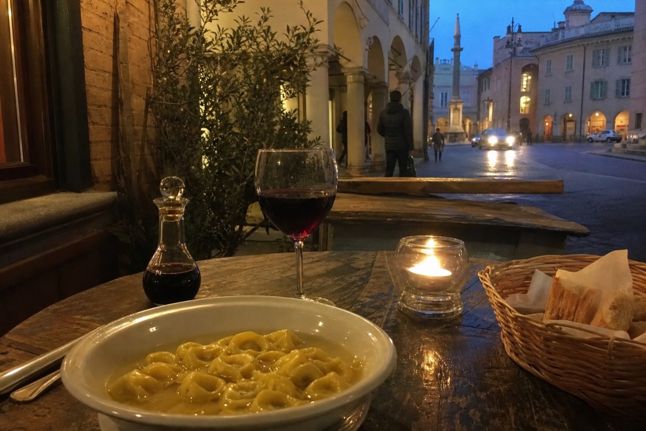 A wooden table set with a plate of tortellini in brodo, a glass of Lambrusco, and a bottle of Traditional Balsamic vinegar